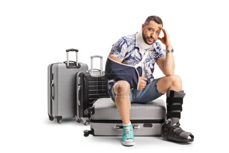 Foto de Turista varón triste con un brazo y una pierna lesionados sentado en maletas aisladas sobre fondo blanco - Imagen libre de derechos