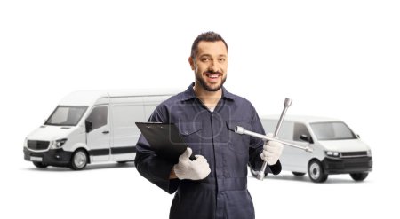 Foto de Mecánico automático sosteniendo un portapapeles y una llave inglesa y posando frente a furgonetas aisladas sobre fondo blanco - Imagen libre de derechos