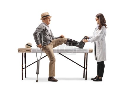 Téléchargez les photos : Homme âgé avec une attelle de jambe assise sur un lit et une femme médecin vérifiant son pied isolé sur fond blanc - en image libre de droit
