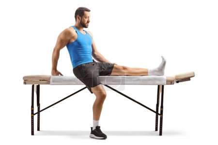 Téléchargez les photos : Athlète masculin avec une jambe blessée assise sur un lit de massage isolé sur fond blanc - en image libre de droit