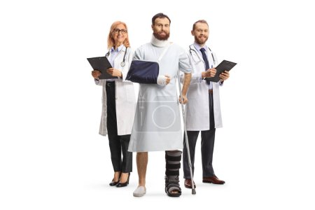 Téléchargez les photos : Équipe de médecins derrière un patient masculin avec une botte orthopédique et un col cervical dans une robe d'hôpital isolé sur fond blanc - en image libre de droit