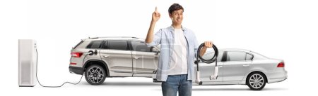 Téléchargez les photos : Homme tenant un câble pour la charge du véhicule électrique et geste pouces vers le haut isolé sur fond blanc - en image libre de droit
