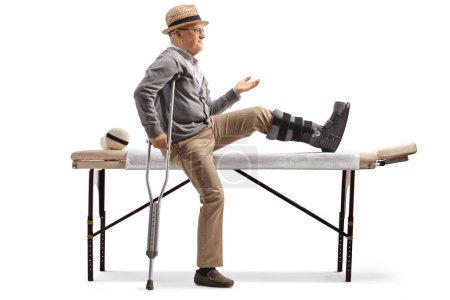 Foto de Hombre mayor con un aparato ortopédico sentado en una cama para fisioterapia aislado sobre fondo blanco - Imagen libre de derechos