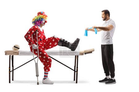 Téléchargez les photos : Physiothérapeute montrant l'exercice à un clown avec une jambe blessée isolé sur fond blanc - en image libre de droit