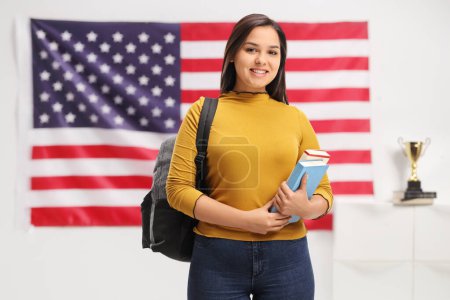 Photo for Female student with a backpack and books smiling in front of an american flag - Royalty Free Image