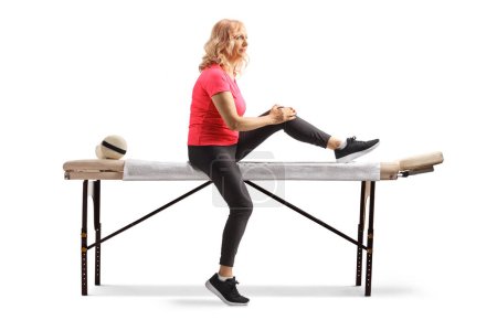 Téléchargez les photos : Femme mûre dans un vêtement de sport assis sur une table de thérapie et tenant son genou isolé sur fond blanc - en image libre de droit