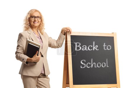 Téléchargez les photos : Professeure tenant des livres et s'appuyant sur un tableau noir avec texte de retour à l'école isolé sur fond blanc - en image libre de droit