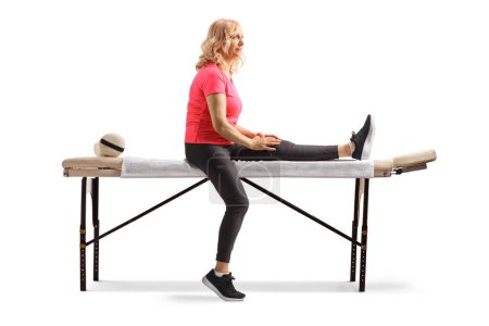 Téléchargez les photos : Femme mûre avec une jambe blessée assise sur une table de thérapie isolée sur fond blanc - en image libre de droit