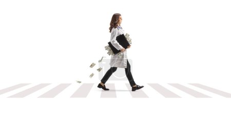 Téléchargez les photos : Jeune femme médecin marchant avec une mallette pleine d'argent à un passage pour piétons isolé sur fond blanc - en image libre de droit