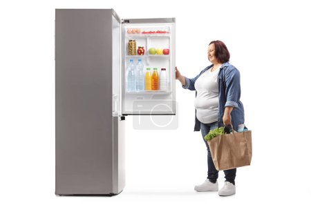 Photo for Overweight woman with grocery bags opening a fridge isolated on white background - Royalty Free Image