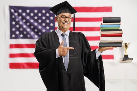 Foto de Hombre maduro en un vestido de graduación sosteniendo una pila de libros y señalando delante de la bandera de EE.UU. - Imagen libre de derechos