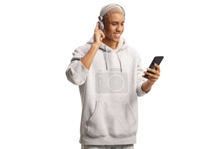 Téléchargez les photos : Jeune homme afro-américain avec écouteurs écoutant de la musique à partir d'un téléphone portable isolé sur fond blanc - en image libre de droit