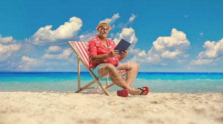 Foto de Turista varón maduro con un libro sentado en una silla bech junto al mar - Imagen libre de derechos