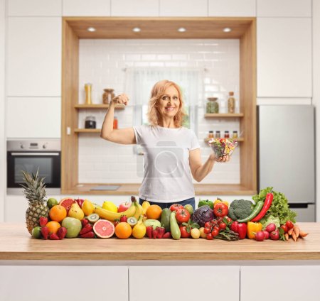 Téléchargez les photos : Heureuse femme forte tenant un bol de salade derrière les fruits et légumes sur un comptoir de cuisine - en image libre de droit