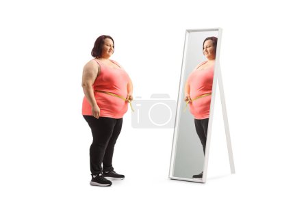 Téléchargez les photos : Jeune femme en surpoids mesurant sa taille devant un miroir isolé sur fond blanc - en image libre de droit