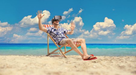Téléchargez les photos : Homme à la plage portant un casque VR et assis sur une chaise longue au bord de la mer - en image libre de droit