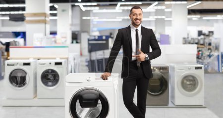 Téléchargez les photos : Jeune homme professionnel dans un magasin appuyé sur une machine à laver et pointant - en image libre de droit