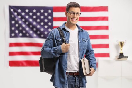 Téléchargez les photos : Étudiant avec un sac à dos et des livres regardant la caméra et souriant devant un drapeau américain - en image libre de droit