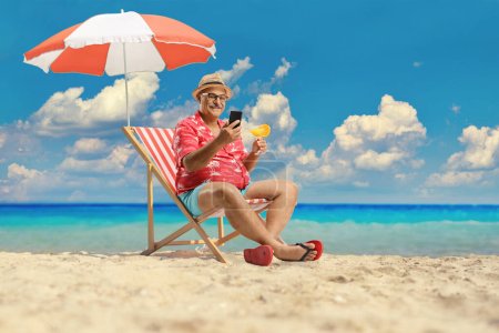 Téléchargez les photos : Homme à la plage assis sur une chaise longue au bord de la mer avec un cocktail et en utilisant un téléphone portable - en image libre de droit