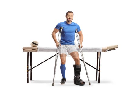 Téléchargez les photos : Joueur de football avec des béquilles assis sur un lit de thérapie physique isolé sur fond blanc - en image libre de droit