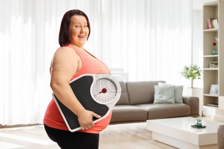 Photo for Smiling chubby woman standing at home and holding a weight scale - Royalty Free Image
