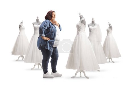 Téléchargez les photos : Femme en surpoids regardant les robes de mariée et la pensée isolée sur fond blanc - en image libre de droit