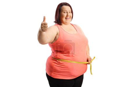 Téléchargez les photos : Femme souriante en surpoids mesurant sa taille et gesticulant pouces vers le haut isolé sur fond blanc - en image libre de droit