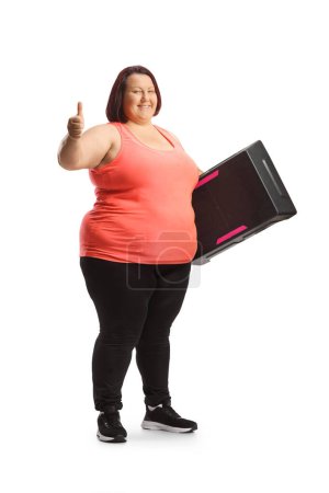 Téléchargez les photos : Plan complet d'une femme corpulente tenant une plate-forme aérobie pas à pas et montrant les pouces isolés sur fond blanc - en image libre de droit