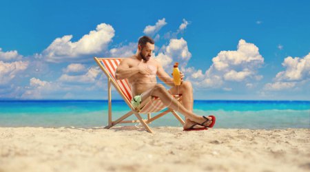 Téléchargez les photos : Homme assis sur une chaise longue et mettant de la crème solaire sur la plage au bord de la mer - en image libre de droit