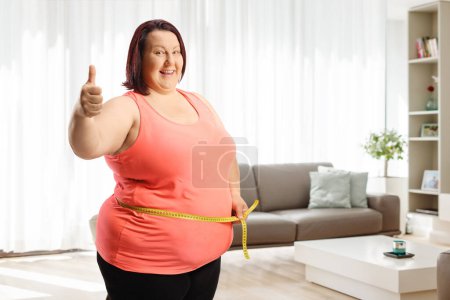 Photo for Happy overweight woman measuring her waist at home and gesturing thumbs up - Royalty Free Image
