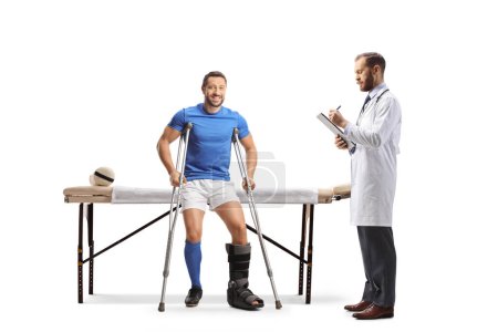 Téléchargez les photos : Joueur de football avec des béquilles assis sur un lit de physiothérapie et un médecin écrivant un document isolé sur fond blanc - en image libre de droit