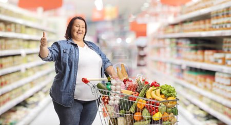 Téléchargez les photos : Femme en surpoids dans un supermarché debout avec un panier et les pouces geste vers le haut - en image libre de droit