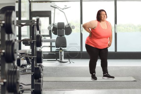 Foto de Mujer joven con sobrepeso sosteniendo su dolorosa espalda en un gimnasio - Imagen libre de derechos