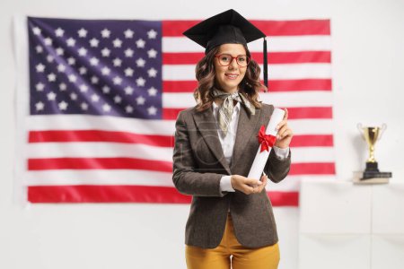 Photo for American female student with a graduation hat holding a diploma in front of a USA flag - Royalty Free Image