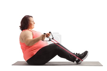 Téléchargez les photos : Plan de profil complet d'une femelle corpulente faisant de l'exercice avec une bande de résistance et assise sur un tapis isolé sur fond blanc - en image libre de droit