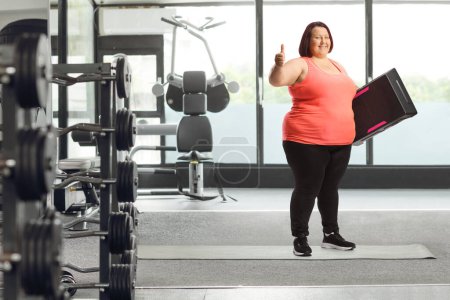 Photo for Overweight woman holding a step aerobic platform  and showing thumbs up at the gym - Royalty Free Image