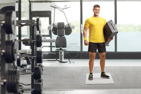 Photo for Full length portrait of a young man holding an aerobic stepper and smiling at the gym - Royalty Free Image