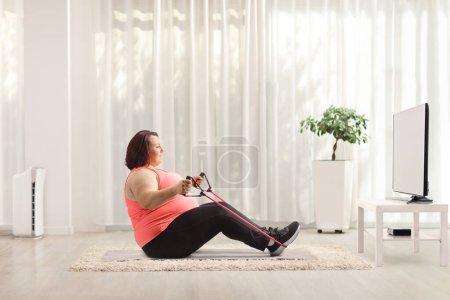 Téléchargez les photos : Plan de profil complet d'une femme corpulente faisant de l'exercice avec une bande de résistance et assise devant la télévision - en image libre de droit