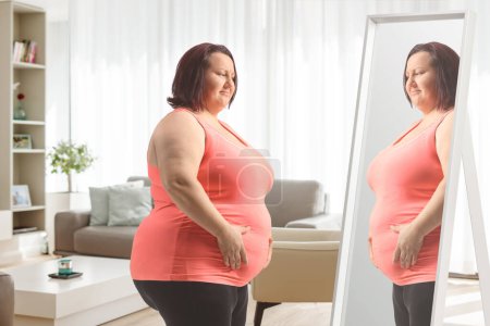 Photo for Overweight woman holding her belly and looking at a mirror at hom - Royalty Free Image