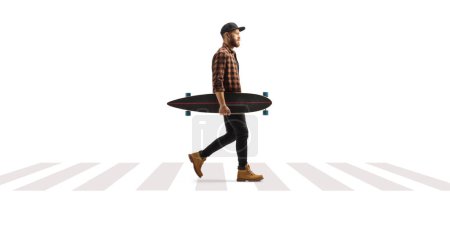 Photo for Full length profile shot of a guy carrying a longboard and walking at a pedestrian crosswalk isolated on white background - Royalty Free Image