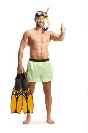Téléchargez les photos : Portrait complet d'un jeune homme en maillot de bain avec un masque de plongée tenant des nageoires de plongée et des pouces gestuels isolés sur fond blanc - en image libre de droit