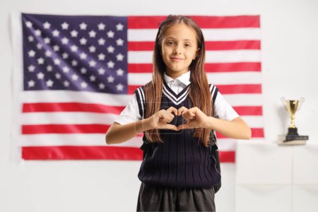 Téléchargez les photos : Portrait d'une écolière faisant signe d'un cœur avec les mains devant un drapeau américain - en image libre de droit