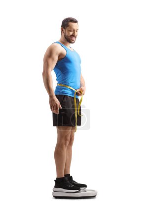 Photo for Full length shot of a fit man in sportswear measuring waist and standing on a weight scale isolated on white background - Royalty Free Image
