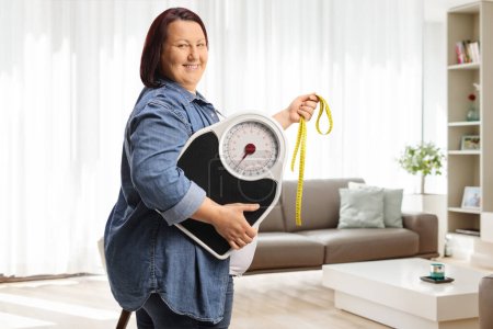 Téléchargez les photos : Sourire femme en surpoids tenant une balance de poids et un ruban à mesurer à la maison - en image libre de droit