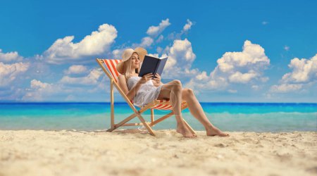 Photo for Young woman in a white dress sitting on a beach chair and reading a book by the sea - Royalty Free Image