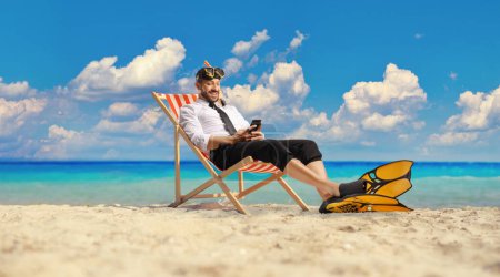 Téléchargez les photos : Businessman with snorkelling fins and mask sitting on a bech chair by the sea and typing on a smartphone - en image libre de droit