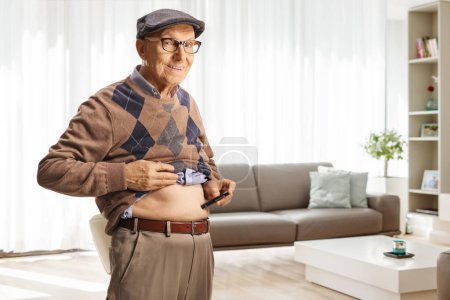 Foto de Elderly man poking abdomen with an insulin pen at home in a living room and looking at camera - Imagen libre de derechos