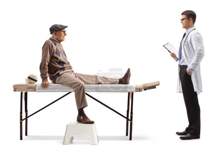 Foto de Elderly man with an injured leg sitting at a physical therapy bed and talking to a doctor isolated on white background - Imagen libre de derechos
