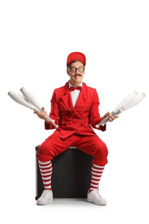 Téléchargez les photos : Interprète drôle en costume rouge assis et tenant des clubs de jonglerie isolés sur fond blanc - en image libre de droit