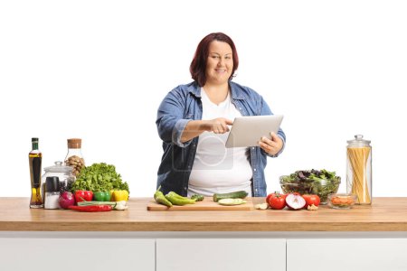 Téléchargez les photos : Jeune femme corpulente tenant une tablette numérique derrière un comptoir de cuisine avec de la nourriture isolée sur fond blanc - en image libre de droit